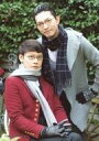 【中古】生写真(男性)/執事歌劇団 環・隈川/膝上・衣装赤・グレー・マフラー・環座り・右向き・両手組み・野外・「執事歌劇団 Autumn Concert」東急ハンズ 花の配剤/執事歌劇団公式生写真【タイムセール】