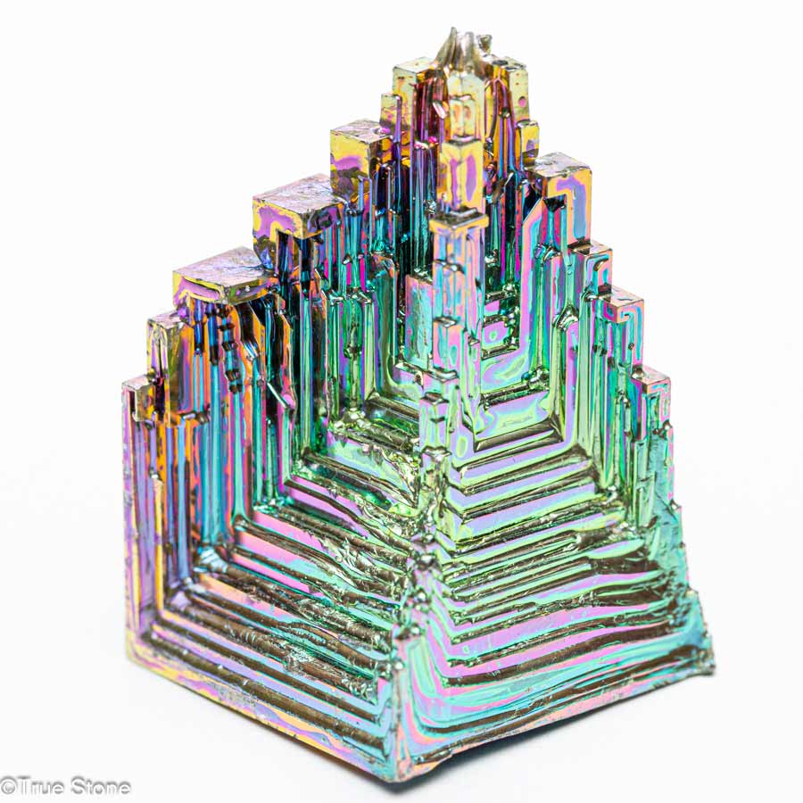 ビスマス結晶"Bismuth crystal"は、その非常に特異な形状と虹色の輝きによって、多くの人々を魅了する金属結晶の一つです。 周期表上では原子番号83、記号Biとして知られ、比較的重い元素に分類されます。 天然にも存在しますが、通常は非常に希少で、私たちが一般に目にするビスマス結晶のほとんどは人工的に作られたものです。 しかし、それが決してその美しさや魅力を損なうものではなく、むしろ人の手によって引き出された構造美が、金属結晶という素材に芸術性をもたらしていると言っても過言ではありません。 ビスマスは低融点金属であり、271.5℃という比較的低い温度で溶ける特性があります。 この性質を利用して、比較的簡単な装置でビスマス結晶を作ることが可能となっており、近年では理科教育や趣味の鉱物収集の一環としても人気があります。 結晶を生成するには、まずビスマスを加熱して完全に溶融させ、その後ゆっくりと冷却する工程を経る必要があります。 この冷却過程で、ビスマス原子が一定の秩序をもって再び固体へと再結晶していく際に、特徴的な「階段状の構造」や「螺旋的な段差模様」を形成します。 これは「ホッパー結晶」とも呼ばれる形態で、成長の速い外縁部に対して中心部の成長が遅いために、空洞状で段差のある立方体や六角形のような幾何学的形状が生まれるのです。 このような独特の結晶構造に加えて、ビスマス結晶の大きな魅力の一つがその表面を彩る虹色の光沢、つまり「干渉色」です。ビスマスそのものの金属色は銀白色に近いのですが、酸化膜が形成されることによって光の干渉現象が起こり、青、緑、紫、赤などのさまざまな色が見えるようになります。 この色彩は結晶ごとに異なり、酸化膜の厚さや角度によって異なるため、まったく同じ色合いの結晶は存在しないという点も、ビスマス結晶の希少性と価値を高めている要因です。 また、これらの干渉色は人工的に制御することもある程度可能であり、結晶生成時の温度管理や冷却速度の調整によって微妙な色の違いを作り出すことができます。 物理的な特徴としては、ビスマスは比較的脆く、衝撃を加えると割れやすい性質があります。 その一方で、他の重金属と比較して毒性が非常に低い、むしろほぼ無毒であることから、化粧品や医薬品にも利用されてきた実績があります。 例えば、胃薬に含まれるビスマスサブサリチル酸塩は、消化器系の保護や殺菌作用があることで知られています。 これにより、ビスマスは「安全な重金属」として注目され、鉱物収集や装飾目的だけでなく、さまざまな産業用途にも応用されています。 さらに興味深いのは、ビスマスが示す特異な物理的性質です。 たとえば、金属でありながら常温で水よりも体積が大きくなる、つまり凝固すると膨張する性質を持っている数少ない物質の一つであることです。 これは鉛やスズなどとは正反対の挙動であり、型に流し込んだビスマスが冷えて固まると、型の隅々まで満たされやすいという利点があります。 こうした性質のため、かつては鉛の代替品として活字の鋳造や金属部品の精密成型などにも使われていました。 科学技術的な側面では、ビスマスは超伝導や熱電材料としての研究対象としても注目されています。 ビスマスを含む一部の合金や化合物は、特定の条件下で電気抵抗がゼロになる超伝導状態を示すことがあり、これは量子力学的な現象として多くの物理学者の関心を集めています。 また、ビスマスは熱電効果、つまり温度差を電気エネルギーに変換する能力に優れており、これを利用した発電デバイスや冷却システムへの応用も模索されています。 一方、芸術的な視点からビスマス結晶を見ると、その人工的に育成された美しさは天然石や鉱物とは異なるユニークな魅力を持ちます。 人間の手によって生まれながらも、自然法則に従って成長する結晶の秩序美は、「人工の自然」とでも呼ぶべき特別な存在感を放っています。 近年では、アクセサリーやインテリアオブジェとしても高い人気を誇り、スチームパンクやサイバーパンクといった美学と融合した作品の素材としても用いられることが増えています。 総括すると、ビスマス結晶は金属元素としての特異な性質、美しい結晶構造と虹色の光沢、さらには安全性と応用可能性を兼ね備えた非常に魅力的な物質です。 科学と芸術、産業と美術という異なる領域にまたがって存在意義を発揮するこの結晶は、単なる観賞用の鉱物にとどまらず、私たちの物質世界と知的好奇心の両方に訴えかける存在と言えるでしょう。 ビスマス（元素番号83）の元々の色はシルバーで最初から虹色に輝く金属ではありません。 ビスマスを全て溶かし階段状に結晶化させた段階で、溶けていないビスマスの液体を取りだします。 すると、ビスマス結晶の表面が空気と反応し酸化膜が形成されます。 これが虹色に輝く秘密です。 ビスマスは低融点の金属（融点約271°C）なのでガスバーナー等で溶かしてビスマス結晶を作ることができます。 ※溶かした時点では非常に高温なので取り扱いには厳重注意です ただし、大きなビスマス結晶を作製するのは至難の業です。 YouTubeの「GENKI LABO」では、サイエンスアーティストの市岡元気さんが巨大ビスマス結晶の作製動画をアップされていますが相当悪戦苦闘されているようです。 色々な方法を考えて巨大ビスマス結晶を作られているようなので今後の進展に期待しています。 ビスマス結晶が虹色に見えるのは、酸化膜による光の干渉が原因です。 1. 酸化膜の形成 ビスマスを溶かして冷却する過程で、表面が空気と反応し「酸化ビスマス（Bi?O?）」の薄い膜が形成されます。 この酸化膜は非常に薄く、厚さがナノメートル（nm）単位です。 2. 光の干渉 酸化膜の厚さが異なると光が反射する際に、異なる波長の光が強め合ったり（強めの干渉）、打ち消し合ったり（弱めの干渉）します。 これにより、見る角度や膜の厚さによってさまざまな「虹色（干渉色）」が生まれるのです。 例 薄い膜 → 青・紫 やや厚い膜 → 緑・黄色 さらに厚い膜 → 赤・オレンジ この現象は、シャボン玉の膜や油膜が張った水たまりが虹色に見えるのと同じ原理です。 3. 色の変化 酸化膜の厚みは冷却速度や温度によって変わるため、ビスマス結晶の色合いは作り方によって異なります。 また、酸化膜を削ったり、化学処理をしたりすることで色を変えることも可能です。 まとめ ビスマスが虹色になるのは、表面にできた酸化膜が光を干渉させることで特定の色を反射するからです。 この現象によって、ビスマス結晶は独特の美しい色合いを持つのです。 レッドツェッペリンのStairway to Heaven（天国への階段）を彷彿されるビスマス人工結晶。 虹の階段を登って行けばそこに天国が存在するような形状です。 ビスマスはもろくて弱い結晶体で破損する恐れがございますのでお取り扱いにはご注意願います。 ビスマス結晶には同じ形・同じ色の物はこの世の中に一切存在しません。 当店のビスマス結晶は全て一点物でお写真に写る商品を発送しています。 「これだ」と思えるビスマス結晶と出会えたならば一生付き合えるお宝となる事でしょう。 【お写真に写っている商品を発送致します】 【サイズ】 ・縦　約41mm ・横　約26.8mm ・厚さ　約26mm 【重量】約62g 「天国への階段」 ビスマスが放つ虹色結晶の真実 True Stone 天然石の真実