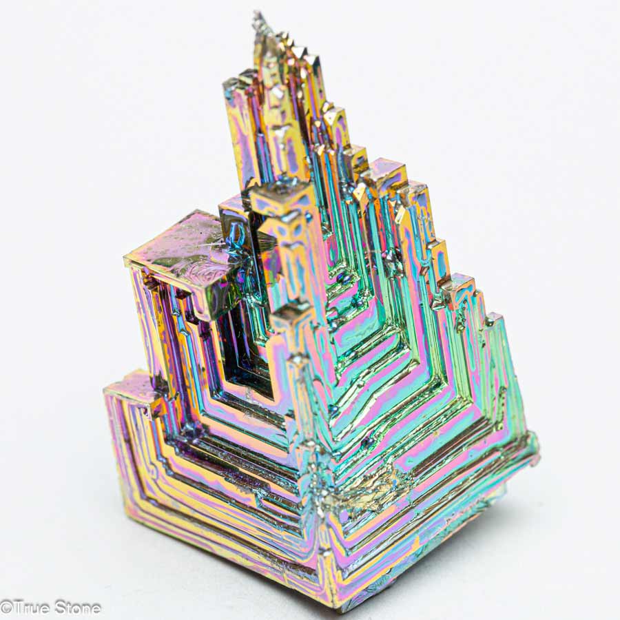 ビスマス結晶"Bismuth crystal"は、その非常に特異な形状と虹色の輝きによって、多くの人々を魅了する金属結晶の一つです。 周期表上では原子番号83、記号Biとして知られ、比較的重い元素に分類されます。 天然にも存在しますが、通常は非常に希少で、私たちが一般に目にするビスマス結晶のほとんどは人工的に作られたものです。 しかし、それが決してその美しさや魅力を損なうものではなく、むしろ人の手によって引き出された構造美が、金属結晶という素材に芸術性をもたらしていると言っても過言ではありません。 ビスマスは低融点金属であり、271.5℃という比較的低い温度で溶ける特性があります。 この性質を利用して、比較的簡単な装置でビスマス結晶を作ることが可能となっており、近年では理科教育や趣味の鉱物収集の一環としても人気があります。 結晶を生成するには、まずビスマスを加熱して完全に溶融させ、その後ゆっくりと冷却する工程を経る必要があります。 この冷却過程で、ビスマス原子が一定の秩序をもって再び固体へと再結晶していく際に、特徴的な「階段状の構造」や「螺旋的な段差模様」を形成します。 これは「ホッパー結晶」とも呼ばれる形態で、成長の速い外縁部に対して中心部の成長が遅いために、空洞状で段差のある立方体や六角形のような幾何学的形状が生まれるのです。 このような独特の結晶構造に加えて、ビスマス結晶の大きな魅力の一つがその表面を彩る虹色の光沢、つまり「干渉色」です。ビスマスそのものの金属色は銀白色に近いのですが、酸化膜が形成されることによって光の干渉現象が起こり、青、緑、紫、赤などのさまざまな色が見えるようになります。 この色彩は結晶ごとに異なり、酸化膜の厚さや角度によって異なるため、まったく同じ色合いの結晶は存在しないという点も、ビスマス結晶の希少性と価値を高めている要因です。 また、これらの干渉色は人工的に制御することもある程度可能であり、結晶生成時の温度管理や冷却速度の調整によって微妙な色の違いを作り出すことができます。 物理的な特徴としては、ビスマスは比較的脆く、衝撃を加えると割れやすい性質があります。 その一方で、他の重金属と比較して毒性が非常に低い、むしろほぼ無毒であることから、化粧品や医薬品にも利用されてきた実績があります。 例えば、胃薬に含まれるビスマスサブサリチル酸塩は、消化器系の保護や殺菌作用があることで知られています。 これにより、ビスマスは「安全な重金属」として注目され、鉱物収集や装飾目的だけでなく、さまざまな産業用途にも応用されています。 さらに興味深いのは、ビスマスが示す特異な物理的性質です。 たとえば、金属でありながら常温で水よりも体積が大きくなる、つまり凝固すると膨張する性質を持っている数少ない物質の一つであることです。 これは鉛やスズなどとは正反対の挙動であり、型に流し込んだビスマスが冷えて固まると、型の隅々まで満たされやすいという利点があります。 こうした性質のため、かつては鉛の代替品として活字の鋳造や金属部品の精密成型などにも使われていました。 科学技術的な側面では、ビスマスは超伝導や熱電材料としての研究対象としても注目されています。 ビスマスを含む一部の合金や化合物は、特定の条件下で電気抵抗がゼロになる超伝導状態を示すことがあり、これは量子力学的な現象として多くの物理学者の関心を集めています。 また、ビスマスは熱電効果、つまり温度差を電気エネルギーに変換する能力に優れており、これを利用した発電デバイスや冷却システムへの応用も模索されています。 一方、芸術的な視点からビスマス結晶を見ると、その人工的に育成された美しさは天然石や鉱物とは異なるユニークな魅力を持ちます。 人間の手によって生まれながらも、自然法則に従って成長する結晶の秩序美は、「人工の自然」とでも呼ぶべき特別な存在感を放っています。 近年では、アクセサリーやインテリアオブジェとしても高い人気を誇り、スチームパンクやサイバーパンクといった美学と融合した作品の素材としても用いられることが増えています。 総括すると、ビスマス結晶は金属元素としての特異な性質、美しい結晶構造と虹色の光沢、さらには安全性と応用可能性を兼ね備えた非常に魅力的な物質です。 科学と芸術、産業と美術という異なる領域にまたがって存在意義を発揮するこの結晶は、単なる観賞用の鉱物にとどまらず、私たちの物質世界と知的好奇心の両方に訴えかける存在と言えるでしょう。 ビスマス（元素番号83）の元々の色はシルバーで最初から虹色に輝く金属ではありません。 ビスマスを全て溶かし階段状に結晶化させた段階で、溶けていないビスマスの液体を取りだします。 すると、ビスマス結晶の表面が空気と反応し酸化膜が形成されます。 これが虹色に輝く秘密です。 ビスマスは低融点の金属（融点約271°C）なのでガスバーナー等で溶かしてビスマス結晶を作ることができます。 ※溶かした時点では非常に高温なので取り扱いには厳重注意です ただし、大きなビスマス結晶を作製するのは至難の業です。 YouTubeの「GENKI LABO」では、サイエンスアーティストの市岡元気さんが巨大ビスマス結晶の作製動画をアップされていますが相当悪戦苦闘されているようです。 色々な方法を考えて巨大ビスマス結晶を作られているようなので今後の進展に期待しています。 ビスマス結晶が虹色に見えるのは、酸化膜による光の干渉が原因です。 1. 酸化膜の形成 ビスマスを溶かして冷却する過程で、表面が空気と反応し「酸化ビスマス（Bi?O?）」の薄い膜が形成されます。 この酸化膜は非常に薄く、厚さがナノメートル（nm）単位です。 2. 光の干渉 酸化膜の厚さが異なると光が反射する際に、異なる波長の光が強め合ったり（強めの干渉）、打ち消し合ったり（弱めの干渉）します。 これにより、見る角度や膜の厚さによってさまざまな「虹色（干渉色）」が生まれるのです。 例 薄い膜 → 青・紫 やや厚い膜 → 緑・黄色 さらに厚い膜 → 赤・オレンジ この現象は、シャボン玉の膜や油膜が張った水たまりが虹色に見えるのと同じ原理です。 3. 色の変化 酸化膜の厚みは冷却速度や温度によって変わるため、ビスマス結晶の色合いは作り方によって異なります。 また、酸化膜を削ったり、化学処理をしたりすることで色を変えることも可能です。 まとめ ビスマスが虹色になるのは、表面にできた酸化膜が光を干渉させることで特定の色を反射するからです。 この現象によって、ビスマス結晶は独特の美しい色合いを持つのです。 レッドツェッペリンのStairway to Heaven（天国への階段）を彷彿されるビスマス人工結晶。 虹の階段を登って行けばそこに天国が存在するような形状です。 ビスマスはもろくて弱い結晶体で破損する恐れがございますのでお取り扱いにはご注意願います。 ビスマス結晶には同じ形・同じ色の物はこの世の中に一切存在しません。 当店のビスマス結晶は全て一点物でお写真に写る商品を発送しています。 「これだ」と思えるビスマス結晶と出会えたならば一生付き合えるお宝となる事でしょう。 【お写真に写っている商品を発送致します】 【サイズ】 ・縦　約44mm ・横　約24mm ・厚さ　約24mm 【重量】約50g 「天国への階段」 ビスマスが放つ虹色結晶の真実 True Stone 天然石の真実