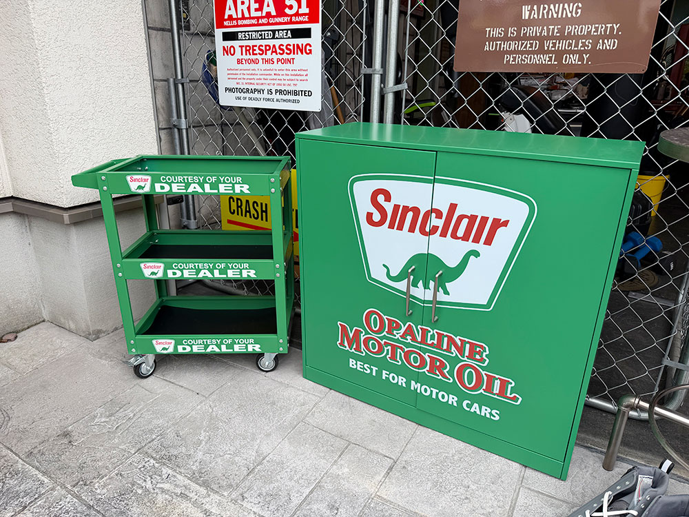 アメリカンガレージを作ってると、 看板を飾って、 オイル缶を並べて、 ネオンサインを灯して・・・・ そのうち欲しくなってくるのが、 本場のショップやサービスステーションにありそうな「什器」なんですよねー そこで見つけたのがコレ！ 恐竜マークでおなじみ、 シンクレアのスチールキャビネット＆キャスター付きスチールワゴンの2点セット！ いやー、 この2台は並べるとメチャクチャ効く！ 何と言っても、 このシンクレアグリーン！ 単品でも十分目立つんだけど、 同じグリーンのキャビネットとワゴンが揃うと、 そこだけ昔のアメリカのサービスステーションみたいな景色になるんです。 まず大きい方が、 シンクレアのスチールキャビネット！ 左右2枚の扉いっぱいに、 あの恐竜マーク入りのSinclairロゴをドーン！ さらに昔のモーターオイル広告を思わせるレタリングが入っていて、 もうこれ、 キャビネットというより「扉の付いた巨大なシンクレア看板」なんですよ！ 何も入れなくても、 置いた瞬間からガレージの主役！ だけど扉を開けると、 中はしっかり収納スペースになっていて、 棚板は高さ調節も可能！ 工具やオイル、 ケミカル用品、 パーツ類、 ヘルメットなんかをまとめて収納できます。 サイズはW79.5cm×D32cm×H78cm。 特に嬉しいのが、 奥行きわずか32cm！ 本場のアンティーク什器って、 見つけてもビックリするくらい高かったり、 「これ、どこに置くねん！」ってくらいデカかったりするんですよねー その点コイツは、 存在感はしっかりありながら、 壁際にも置きやすい！ 日本のガレージにも取り入れやすいサイズ感ってところがイイんです。 そしてその横に並ぶのが、 シンクレアのキャスター付きスチールワゴン！ こちらはW50cm×D30cm×H67.5cmの3段式！ フロントからサイドまで、 シンクレアらしいモーターオイルの広告デザインが入っているので、 どこから見てもちゃんとシンクレア！ 壁際に置いておくだけじゃなく、 ガレージの真ん中へ引っ張り出した時まで、 ちゃんと絵になるんです。 工具やスプレー缶、 オイルやケミカル用品を載せて、 作業する時は愛車の横までゴロゴロッ！ サイドにはハンドルも付いているので、 必要な物を載せたままワゴンごと移動できます。 そしてこの2台をセットにした一番の理由がココ！ キャビネットは「収納する基地」。 ワゴンは「動かして使う相棒」。 普段使わない工具やストック品はキャビネットへ。 今日使う物だけワゴンへ載せて、 愛車の横までゴロゴロッ！ 作業が終わったら、 またキャビネットの横へ戻す。 見た目が揃うだけじゃなく、 2台揃うことでちゃんと使いやすいんです。 もちろん工具だけじゃなく、 アメリカ雑貨のディスプレイ什器にするのもオススメ！ キャビネットの天板にはオイル缶や広告キャラクター。 ワゴンにはマグやボトル、 ミニカーなんかをズラズラッ！ 多少ゴチャッと置いても、 このシンクレアグリーンのおかげで、 そのゴチャゴチャ感までアメリカのショップっぽい景色に見えてくるから不思議なんですよねー そしてこのシリーズについて、 ひとつ知っておいて欲しいところがあります。 実際に組み立ててみると、 扉の建て付けが少しラフだったり、 左右の扉にまたがるプリントにズレがあったり、 小キズやスレが見られたりと、 日本製家具のような精密な仕上がりではありません。 キッチリとした家具としての完成度を最優先する人には、 正直向いてないと思います。 だけど・・・・ 実際に2台並べてみると、 「いや、それを差し引いてもこの景色は欲しいわ！」 ボクらは完全にそっちでした（笑） 小さなアメリカ雑貨をいっぱい集めるのも楽しいけど、 こういう大物什器がひとつ入ると、 ガレージの景色が一気に変わる！ ましてや今回は、 キャビネットとワゴンの2台揃い！ 家具を2個置くんじゃない。 シンクレアのガレージシーンを丸ごと作る！ キャビネットをドン！ その横へワゴンをゴロゴロッ！ この2台が並んだ姿を見てもらえれば、 セットにしたくなった理由、きっとわかってもらえると思います！（￣ー￣）ニヤリ☆ 【スチールキャビネット】 サイズ：横79.5cm×奥行き32cm×高さ78cm（約） 材質：スチール ※組立式です。 【知っておいていただきたいこと】 ペラペラ感はないですが、全体的にラフな作りです。扉の建付の悪さや、正面ロゴの左右プリントズレ、小キズ、汚れなどは在庫分すべてに見られますので製品仕様となります。予めご了承願います。 誠に恐れ入りますが、沖縄・離島へは配送できません。 【スチールワゴン】 サイズ：横30.5cm×奥行き50cm×高さ67.5cm（約） 材質：スチール ※組み立て式 【知っておいていただきたいこと】 製造工程上、プリント部分のわずかなズレ、微細な擦れや小キズ等が見られる場合がございますが製品仕様となります。予めご了承願います。 誠に恐れ入りますが、沖縄・離島へは配送できません。類似商品はこちらチャンピオン　スチールキャビネット　＆　スチー50,600円シンクレア　スチールキャビネット　※沖縄・離島30,800円シンクレア　スチールワゴン　　ツールワゴン　※17,820円シンクレア　スチールワゴン　　ツールワゴン　ア19,800円ペンズオイル　スチールキャビネット　＆　スチー50,600円ペンズオイル　スチールワゴン　　ツールワゴン　17,820円チャンピオン　スチールワゴン　　ツールワゴン　17,820円チャンピオン　スチールキャビネット　※沖縄・離30,800円ペンズオイル　スチールキャビネット　※沖縄・離30,800円新着商品はこちら2026/10/17ポストジェネラル　ライトウエイト　アイアン　サ5,280円2026/10/17ポストジェネラル　ライトウェイト　アイアンメッ20,900円2026/10/17ポストジェネラル　ライトウェイト　アイアンメッ20,900円～2026/10/20 更新