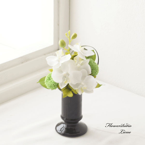 商品情報使用花材◎プリザーブドフラワー　ピンポンマム、あじさい、ミスカンサス、モス◎アーティフィッシャルフラワー　胡蝶蘭ミディ◎花器　ツヤのある茶色の陶器サイズ高さ約19センチメートル横約11センチメートル奥行約12センチメートルラッピングクリアボックスに黒いリボンをお掛けいたします。メッセージカード無料でお付けいたします。内容は備考欄にご記入ください。熨斗必要な方はオプションにて選択お願いします。お名前は備考欄にご記入ください。注意事項◎プリザーブドフラワーの特性上、配送中に葉、実の一部が落ちてしまう事がございます。予めご了承ください。◎ナチュラル素材を使用して一つ一つお作りしております。若干、写真と異なる場合がございますことをご了解ください。◎仕入れの状況により、多少花材が変更になる場合がございます。できる限り、雰囲気を変えずに制作いたしますので、予めご了承ください。お手入れ方法◎水やりは必要ございません。◎直射日光と高温多湿を避けた場所でご使用ください。◎エアコンの風が直接当たる場所でのご使用は、お花のひび割れの原因になります。◎ほこりがついた場合は、冷風のドライアーでほこりを飛ばしてください。◎お洋服や壁、床、などにお花が直接触れると色移りする場合がありますのでご注意ください。胡蝶蘭ミディとグリーンマムの仏花 Sサイズ 花器に固定したタイプです お供え 供養 プリザーブドフラワー 枯れないお花 小さい仏花 納骨堂 オシャレな仏壇に 現代のモダンなお仏壇や納骨堂へのお供えにオススメです こちらはプリザーブドフラワーとアーティフィッシャルフラワーでお作りした仏花です。花器にアレンジした（固定した）タイプです。お手入れのいらないプリザーブドフラワーの仏花は、お仏壇を守るシルバー世代の方に大変喜ばれています。生花のもちが悪い夏場など、一つお持ちになると、どうしてもお花を買いに行けない時などお花を絶やすことがありません。ご自宅用にも贈り物にも大変喜ばれています。花材を吟味して質の良いものだけを使用しておりますので、生花と見間違えるほどの仕上がりです。棘（トゲ）の無いお花を選んで制作しておりますので、贈り物にも安心です。※仕入れの状況により、多少花材が変更になる場合がございます。雰囲気を変えずに制作いたしますので、ご了承ください。 7