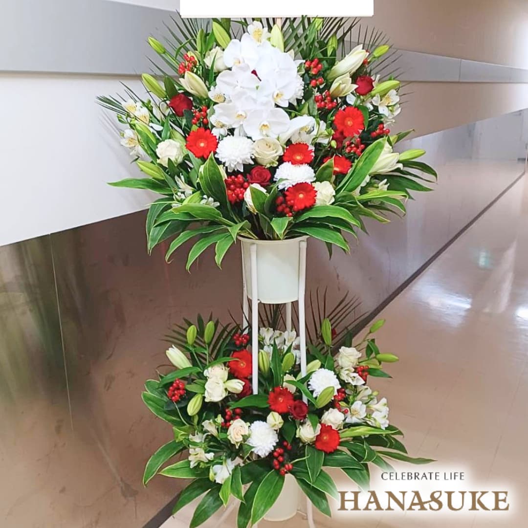 お届け商品 スタンド花 2段 お届け日 仕入れの関係上、1週間ほどお日にちをいただいております。お急ぎの場合は事前にご連絡ください。 使用花材 季節の花をメインに胡蝶蘭を入れておまかせで制作いたします。(胡蝶蘭の数はおまかせになります。) サイズ 幅約100cm　x　高さ約180cm　あくまでも目安です。制作内容により異なります。 お届け内容 オアシス入り専用バケツにスタンド台付き。バケツ部とスタンド台は後日花屋が回収に伺います。 配送方法 全国の優良花屋から手渡しでお届けします。ただし、当店のネットワーク基準に合った花屋のない地域はお届けできない場合がございます。 送料 花助は全国の都市部にある花屋を厳選したフラワーネットワークになります。 都市部であれば送料無料でお届け可能です。 ただ、都市部から離れた郊外へお届けの際は【配送料1,650円（税込）】がかかる場合があります。 ご注文後、配送料については電話もしくはメールにてその旨ご連絡させていただきますのでご安心ください。 無料サービス スタンド花の設置・回収 / 名札 / 配達完了報告 / 画像報告(※画像報告は希望者のみ、配達後5日程度) / 見積書・請求書・領収書全国の花贈りに精通したフラワーコンシェルジュが、お客様一人ひとりのご用途に合った、 満足度の高い花屋をその都度厳選し、最高のフラワーギフトを提供します。