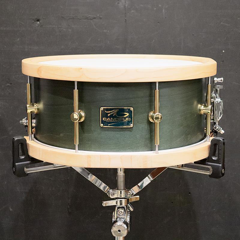■商品紹介＜MO Snare Drum w/Wood Hoop＞厚手のメイプルシェルは潜在的なパワーは持っていますが、鳴らしきることが難しく、ともするとアタック音ばかり目立つ薄っぺらいサウンドになりがちです。しかしこのMOは的確なベアリングエッジを施し、オイルフィニッシュで極薄の塗膜に仕上げることにより、シェルの持つパワーを効率良く引き出すことができます。また、高品質なアメリカンメイプルシェルを使用しつつも、オイルフィニッシュを採用することで材料コストの削減や生産効率の向上をはかり、そのサウンドからは想像もできない驚異的なコストパフォーマンスを実現しています。標準装備のダイキャストフープに比べ、質量の軽量なウッドフープによる「オープンでナチュラル」なサスティーンを加味し、さらに、同素材の部分が多く振動効率が向上しており、サウンドのまとまりをも獲得しました。加えて、オイルフィニッシュされたウッドフープとシェルの、ルックス的なマッチングも非常に良く、ウッドスネアとしての風格と新しい潮流を感じさせる仕上がりとなっております。＜スペック＞サイズ：14''×6.5''シェル：10ply Maple 8mmフィニッシュ：Black Olive Oilラグ：ブラス・チューブフープ：Maple Wood Hoop (Made by CANOPUS) / 8テンションスネアワイヤー：Vintage Snare Wire（CPSL-14DR）付属品：チューニングキー※店頭にて試打を解禁しております為、ウッドフープに打痕や汚れが発生する可能性がございます。予めご了承ください。検索キーワード：イケベカテゴリ_ドラム_スネアドラム_CANOPUS_新品 SW_CANOPUS_新品 JAN:4511239020296 登録日:2026/07/29 スネア スネアドラム カノウプス カノープス