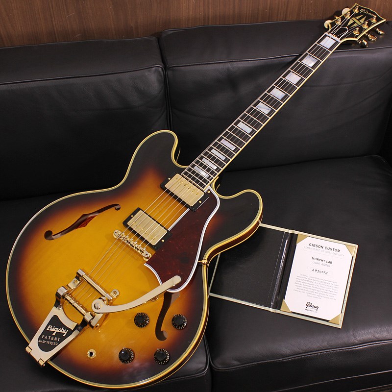 楽器種別：新品エレキギター/Gibson 商品一覧＞＞エレキギター/Gibson/Custom Shop 商品一覧＞＞エレキギター【755，000円〜1，415，000円】 商品一覧＞＞当店のギターは、出荷前に専門スタッフが検品・調整しているので安心です！Gibson Murphy Lab 1959 ES-355 Reissue w/Bigsby Vintage Wide Burst Light Aged SN. A930772【TOTE BAG PRESENT CAMPAIGN】商品説明イケベオーダーのES-355 Bigsbyが極少本数で入荷！只でさえゴージャスな雰囲気のES-355に、レギュラーカラーには無いVintage Wide Burstをセレクトすることでさらに高級アーチトップギターのような気品に満ちた貫禄のルックスを獲得！この美しいギターが手に入るのはイケベだけです！ぜひお見逃しなく！〜 Murphy Lab 〜カスタムショップ内に誕生したMurphy Lab（マーフィー・ラボ）は、ナッシュヴィルのギブソン・カスタムショップ内に新設されたラボで、科学的検証に基づく最新の技術を駆使して、ギターのエイジング（ヴィンテージ・ギターの経時変化した外観と弾き心地を再現する処理）を専門に行います。ギブソン・カスタムショップの立ち上げ時のメンバーであり、ヒストリック・コレクション開発のプロジェクト・リーダーとして尽力したトム・マーフィーは、その後新品のギターにヴィンテージのルックスとフィーリングをもたらすエイジングというテクニックで活躍してきました。トムのヴィンテージ・ギターへの情熱と深い知識に裏打ちされた技術によって、ギブソン・ギターの自然なラッカーのクラック、演奏による摩耗を見事に再現しています。トム・マーフィーとギブソンは、経時変化によりウェザーチェックと呼ばれるクラックを生じさせる、1950年代、1960年代のニトロセルロースラッカーを科学的に解析し、その変化のプロセスをひも解き、あらゆるモデルのプロトタイプによるテストを重ねて、ヴィンテージ同様のナチュラル・ウェザーチェックを再現するラッカー・プロセスを開発しました。マーフィー・ラボは、トム・マーフィーがギブソン・カスタムショップのチームを率いて、プロトタイプをもとに、あらゆるボディシェイプ、フィニッシュ、エイジング・レベルの検証を始めたことから、その構想が生まれました。その結果、これまでにないリアリティとこだわりを追求したエイジングを行うために、ギブソン・カスタムショップ内に設立されました。トム・マーフィーの存在と、その芸術的な作品の数々は、ギブソンの歴史の中で重要な役割を果たし、今もそしてこれからもマーフィー・ラボとして永遠に引き継がれていきます。本機は4段階で設けられた中で2番目に軽度なエイジングレベルとなる「Light Aged」が施された1959 ES-355 Reissueです。ES-355はLes Paulで言うところのアップグレードモデルである「Les Paul Custom」的な位置付けとなり、上級機種特有の豪華な装飾を採用している点が特徴です。ヘッドトップに輝くスプリットダイアモンドインレイ、輪郭をシャープに引き締めるマルチプライ・セルバインディング、オールゴールドのハードウェアに漆黒のエボニー指板の採用等、ES-355ならではの豪華さと圧倒的に重厚な存在感がES-335とは一線を画す雰囲気を醸し出しております。センターブロック同時期のES-335と同様にウインドウ無しのドリルホールを採用しています。Light Agedは軽度の凹みや塗装剥がれ等のダメージ、リアルな塗装の色艶とウェザーチェックを表現。指板エッジにはロールド加工が施され、優れたグリップ感がプレイヤーの皆様にはさらに嬉しい仕上げとなっております。基本的なスペックは最新のHistoric Specを採用。アンポッテッドのCustombucker Alnico IIIピックアップやアップデートされたアッセンブリ、True Historicパーツが引き続き装備されております。ピッキングに鋭く追従する鋭敏なレスポンスは、ギターをアンプにプラグインしたその刹那からプレイヤーを魔法の様にGibsonトーンの虜にしてしまいます！トーンを絞った際の音抜け感の良さは勿論のこと、ヴォリュームを絞っても程良くブライトさがキープされるコントローラブルなアッセンブリーは現代的な使用方法とも素晴らしい相性を見せてくれるでしょう！！原点に回帰することでさらなる進化を続けるGibson Custom Shopの最新型の入荷です。Custom Shop製ならではの素晴らしいトーン、Murphy Labが生み出す迫力満点のエイジングをぜひご堪能下さい！Serial No. A930772 Weight≒4.05kg専用ブラウンハードケース付属商品詳細Body: 3-Ply Maple/Poplar/MapleNeck: Solid Mahogany with long neck tenonFingerboard: EbonyFrets: 22， Authentic Medium-JumboScale Length: 24.75inchNut Width: 1-11/16inchBridge: ABR-1 / Bigsby B7Machine Head: Grover MilkbottleHardware: GoldPickups: Custombucker Alnico III UnpottedControls: 2 Volume 、2 Tone 、3-way selector switchイケベカテゴリ_ギター_エレキギター_Gibson_Custom Shop_新品 JAN:4570155998867 登録日:2023/11/02 エレキギター ギブソン カスタムショップ