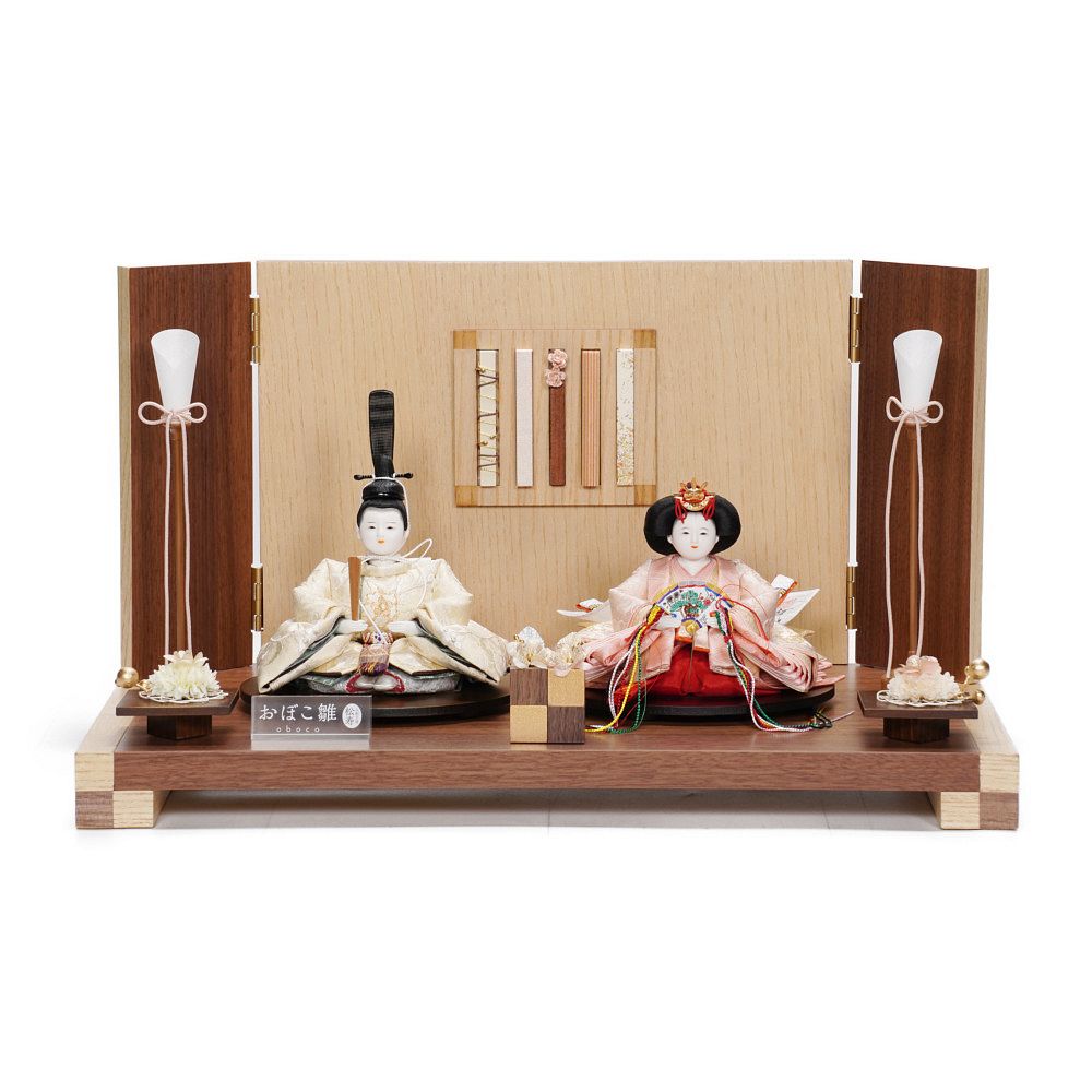【製作】小出松寿 【頭師】市川伯英おぼこ 【規格】三寸おぼこ親王 【仕様】桐木胴、絵羽合わせ、木手 【装飾】木目飾り台、スティック付き木目屏風、油灯、桜、口花、毛氈 【寸法】幅50×奥行30×高さ28(cm) ※手作り品につき仕様が変更になる場合がございます。予めご了承くださいませ。 ※お使いのモニターの設定や発色により、実際の商品と色味が異なって見える場合がございます。 温かみのあるウォールナットを基調とし、表情の異なる五種類のスティックがアクセントになったモダンな飾りです。 真ん丸なお子さんのお顔を表現した、可愛らしいおぼこ雛。 正倉院裂などの上代裂に遺る唐花文は、唐の時代に最も華麗な様相を見せました。 理想化した花を真上から見た形に様式化されています。 優美な意匠のお衣裳で、慶賀の特を華やかに彩ります。 -----------松寿の4つのこだわり---------- 1.【桐木胴(きりもくどう)】 松寿の雛人形は、防虫効果のある樟(くすのき…樟脳の原料)をはさんだ「桐木胴」を使用しています。中心に藺草(いぐさ)を使い安定感のある太串の頭に対応しています。 胴の型部分には針金を埋め込み人形そのものの型崩れを防止しています。 ウレタン胴は虫がつかず長年経っても型が崩れず樟脳などから発生する溶解の心配がないことから、胸元や足元に使用しています。 【特許 第4389193号】 2.【絵羽あわせ(えばあわせ)】 絵羽あわせとは着物の柄合わせのことをいいます。松寿では殿の胸元の柄を合わせて裁断し、仕立てています。生地の用尺(必要m数)もその分余計にかかりますが、装束の柄の美しさを効果的に演出します。 3.【丁寧な縫製】 製品になった時の仕上がりが、いかにきれいに見えるかを常に考え、裁断した生地を一つ一つ丁寧に縫製しています。 4.【バランスの取れた着付け】 着付けで殿は「風格・威厳」、姫は女性らしい「やさしさ」を表現します。頭の挿し方、腕折り(かいなおり)など、長年培われた技術と感性によってなされるものであり、その全てが揃っているのが松寿の「雛人形」です。