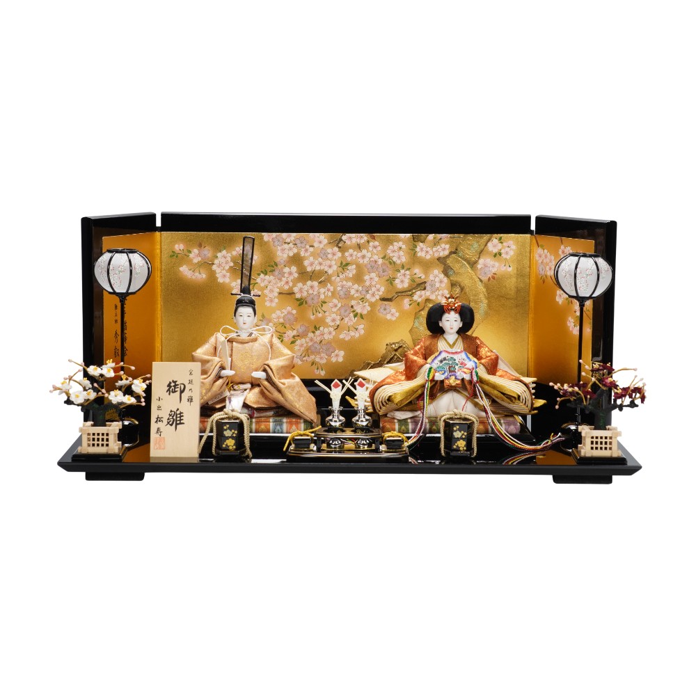 【製作】小出松寿 【頭師】市川伯英 【規格】京十二番親王 【仕様】桐木胴、絵羽合わせ、木手 【装飾】飾り台、屏風、灯り、御花、前飾り、毛氈 【寸法】幅60×奥行35×高さ27(cm) ※手作り品につき仕様が変更になる場合がございます。予めご了承くださいませ。 ※お使いのモニターの設定や発色により、実際の商品と色味が異なって見える場合がございます。 〈本金手描蒔絵 金沢桜華〉 雛具師「秀叡」による、内閣総理大臣賞受賞作品。 金箔の最高峰といわれる「金沢箔」を贅沢に使用した屏風です。 雅やかに輝く金色のキャンパスいっぱいに、手描き蒔絵の桜花が咲き誇っています。 〈牡丹唐草文様〉 牡丹の花の周囲を唐草風の葉でつないだ古典的な意匠です。 牡丹は、あでやかな大輪の花を咲かせることから「百花の王」といわれ、富貴の象徴として親しまれてきました。 唐草は絡みまつわる草の文様で、連続的な形状と厳しい環境下でも生育する強い生命力から、永続的な繁栄を表しています。 最高級の香木である伽羅に由来する「伽羅色」と、赤みを帯びた茶色が味わい深い「弁柄色」のお衣裳の気品あるお雛さまです。 -----------松寿の4つのこだわり---------- 1.【桐木胴(きりもくどう)】 松寿の雛人形は、防虫効果のある樟(くすのき…樟脳の原料)をはさんだ「桐木胴」を使用しています。中心に藺草(いぐさ)を使い安定感のある太串の頭に対応しています。 胴の型部分には針金を埋め込み人形そのものの型崩れを防止しています。 ウレタン胴は虫がつかず長年経っても型が崩れず樟脳などから発生する溶解の心配がないことから、胸元や足元に使用しています。 【特許 第4389193号】 2.【絵羽あわせ(えばあわせ)】 絵羽あわせとは着物の柄合わせのことをいいます。松寿では殿の胸元の柄を合わせて裁断し、仕立てています。生地の用尺(必要m数)もその分余計にかかりますが、装束の柄の美しさを効果的に演出します。 3.【丁寧な縫製】 製品になった時の仕上がりが、いかにきれいに見えるかを常に考え、裁断した生地を一つ一つ丁寧に縫製しています。 4.【バランスの取れた着付け】 着付けで殿は「風格・威厳」、姫は女性らしい「やさしさ」を表現します。頭の挿し方、腕折り(かいなおり)など、長年培われた技術と感性によってなされるものであり、その全てが揃っているのが松寿の「雛人形」です。