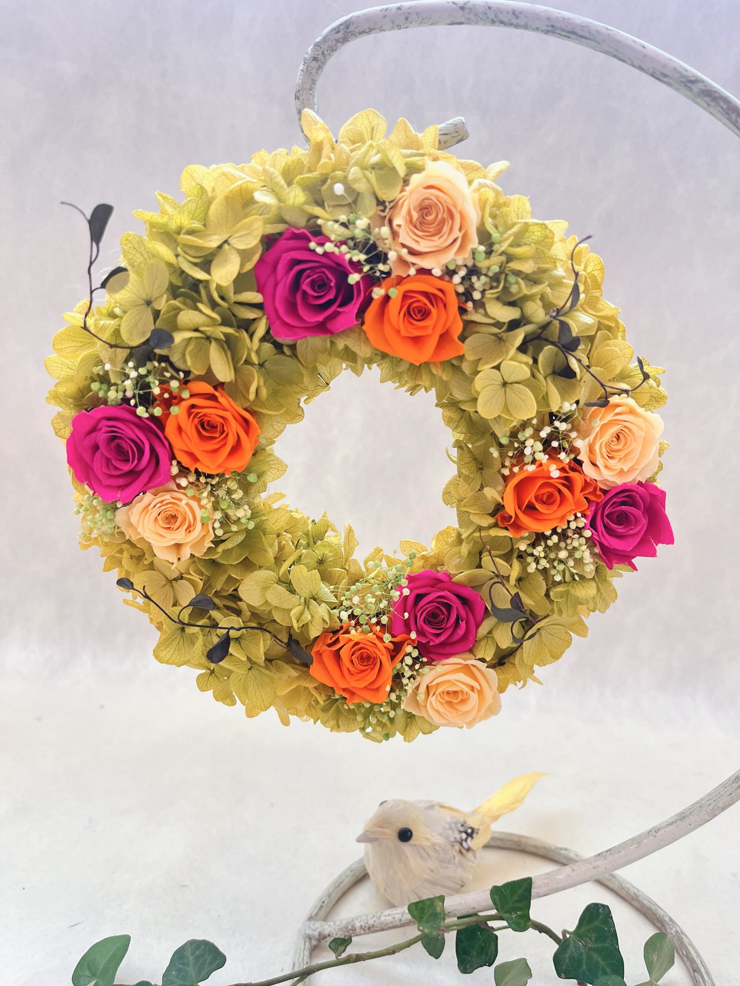 ピンク、オレンジ、黄色の鮮やかなバラのリースです。 ビタミンカラーがいっぱいで、見るだけで元気になれそう！お誕生日や結婚祝い、母の日、敬老の日など、さまざまなお祝いのシーンにぴったりです。お部屋に飾れば、明るく華やかな雰囲気を演出してくれます。特別な贈り物として、大切な人に喜ばれること間違いなしです！ 木製アレンジスタンドに飾ると、お洒落感アップ!! 使用花材:プリザーブドフラワー( バラ　かすみ草　アジサイ)　 サイズ: 直系18センチ プリザーブドフラワーですので水をかけないで下さい。 ※撮影用小物は含まれていません。 木製アレンジスタンドはこちらからご注文下さい。