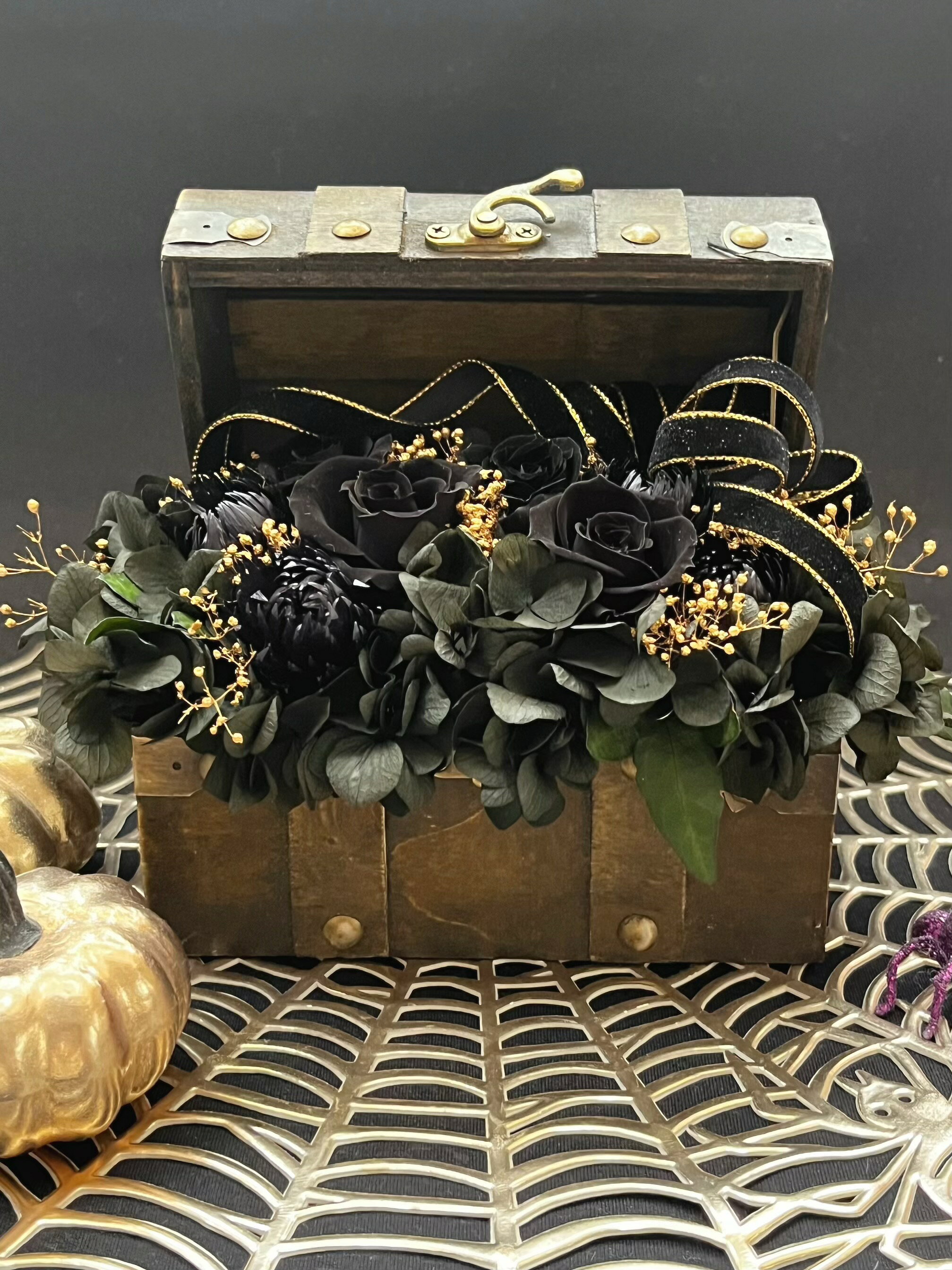黒バラと黒い紫陽花、黒いシルバーデージーを用いたゴージャスな宝箱アレンジです。 金の縁取りが施されたベルベットリボンとゴールドのかすみ草が、全体にきらびやかな印象を与えます。ハロウィンの飾りとして最適で、特別な日のギフトやプレゼントにもぴったりです。誕生日や退職祝いなど、さまざまなシーンで華やかさを演出します。 花材:プリザーブドフラワー(バラ　かすみ草　紫陽花　シルバーデージー　アイビー) トランク:木材 サイズ:横15×13 高さ16センチ(ふたを開けた状態) プリザーブドフラワーですので水をかけないで下さい。 撮影用小物は価格に含まれていません。