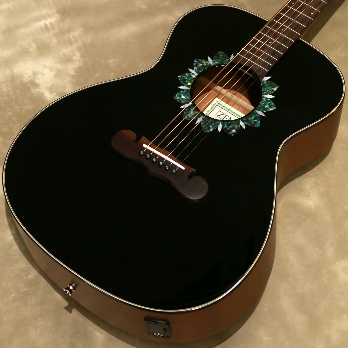 ゼマイティス・アコースティックよりヒマワリをイメージしたロゼッタにラウンド・サウンドホールを採用したグランド・オーディトリアム・サイズにアンダーサドル・ピックアップを搭載したエレクトリック・アコースティック・モデルのCAG-90Fが登場しました。 CAG-90Fは、マザー・オブ・パールのダイヤモンド・インレイが輝く特徴的なヘッドストックを持ったマホガニー・ネックに、ローズウッド・フィンガーボードを採用した20フレット仕様の14フレット・ジョイントとなっています。 ラウンド・サウンドホールの周縁にヒマワリをイメージしたロゼッタ、マスターシュ・ブリッジのオリジナル・デザインは、非常に魅力的です。 トップ・ブレイシングはサウンドホールの形状やボディーのサイズを考慮して、最大限の鳴りを引き出すためにフォワードXブレイシングを採用しています。 ボディー・トップは板材の時に真空状態で熱を加えることで余分な水分やミネラルを取り除いたソリッド・スプルースを使用し、サイドとバックにはサペリを用いて狂いの出にくい安定したボディー剛性を実現しました。 ヘッドストックの突板にはローズウッドを使用し、ペグのボタンはパーロイドを採用しています。 アンダーサドル・ピックアップを搭載し、ボディー・サイドには4バンドEQとクロマティック・チューナー機能を持ったプリアンプを搭載し、お好みのトーンでアンプなどの外部機器に出力できます。 グロス・ブラックのトップにアバロン柄とマザー・オブ・パール柄のロゼッタをラインナップしています。 ●アンダーサドル・ピエゾ・ピックアップからのサウンドをコントロールするプリアンプ・システム。コントロールはVolume、Treble、Middle、Bass、Presence。便利なチューナー機能も装備。9Vバッテリーを使用。 ●その形状から、英語で口ひげを意味する「マスターシュ」と命名されたブリッジ。 ●マザー・オブ・パールのZロゴが入ったヘッドストック。 ●付属ギグバッグは、大きな前面ポケット付き。ダブル・ショルダー・ストラップでバックパック・スタイルでの運搬が可能。 ■Top: Solid Spruce ■Back & Sides: Laminated Sapele ■Bracing: Forward X ■Neck: Mahogany ■Nut Width: 43mm ■Fingerboard & Bridge: Rosewood ■Fretboard Radius: 406mm ■Nut & Saddle: Bone ■Scale Length: 648mm ■Frets: 20 ■Headstock Veneer: Rosewood ■Finish: Urethane ■Body Length: 504mm ■Body Width (Upper Bout): 293mm ■Body Width (LowerBout): 402mm ■Body Depth (Upper): 96mm ■Body Depth (Lower): 120mm ■Preamp: EX-4 with Zemaitis Logo ■Strings: .012 / .016 / .024 / .032 / .042 / .053 [Coated Phosphor Bronze] ■Case: Gigbag ■Weight: 2.25kg