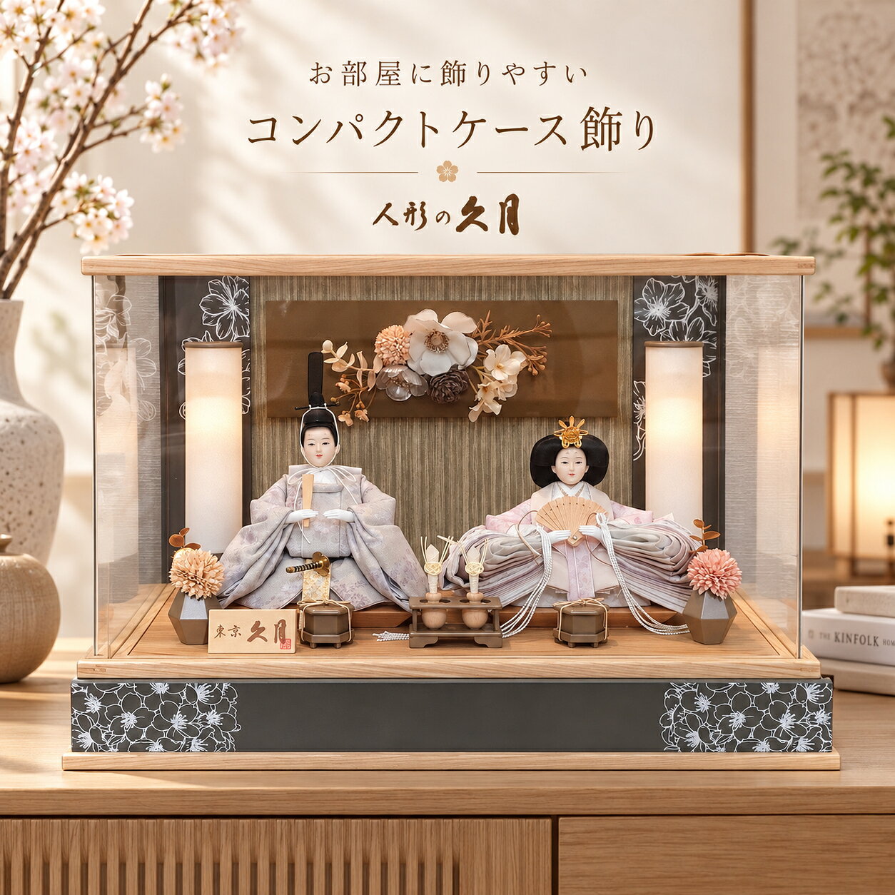 春のお部屋に、やさしい華やぎを。 淡い色彩と灯りがともる雪洞が美しい、久月のアクリルケース親王飾り。 穏やかなお顔立ちのお殿様とお姫様を、白・淡い灰色・やわらかな桃色を基調とした衣裳で上品に仕上げました。 明るい木目と落ち着いた花柄のモダンなケースで、和室にも洋室にも自然になじみます。 人形の久月 正面左下には「東京 久月」の木札を配置。初節句にふさわしい品格を添えます。 アクリルケース入り 大切なお人形をほこりや汚れから守りやすく、飾りやすさにも配慮されています。 奥行22cm チェストやリビングボードなどにも置きやすいスリムな奥行きです。 雪洞が点灯 両脇の雪洞に灯りがともり、夜には温かな光で雛飾りを演出します。 飾った時のサイズ 幅53cm × 奥行22cm × 高さ36cm 大切なお子様の初節句に 女の子の初節句、桃の節句、お孫様への節句祝いにもおすすめです。大切な初節句を、久月の雛人形とともに。 お子様の健やかな成長と幸せを願う桃の節句。 雛人形を飾り、ご家族でお祝いする時間そのものが、かけがえのない思い出になります。 現代のお部屋にも飾りやすい 幅53cmの華やかさを持ちながら奥行きは22cm。リビングボードやチェストにも置きやすいサイズです。 灯りがともる雪洞 温かな光がお殿様とお姫様を照らし、夜には昼とは異なる表情を楽しめます。 お孫様への初節句のお祝いにもおすすめです。
