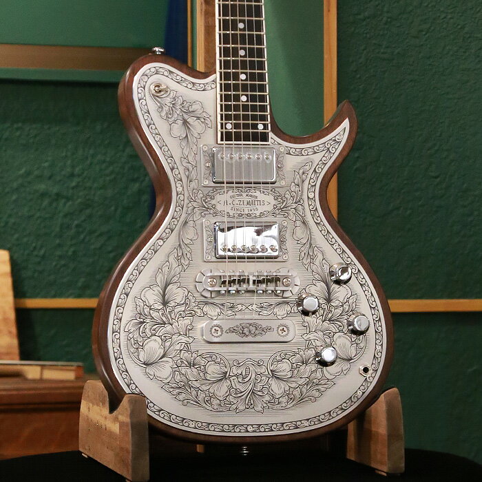 ゼマイティスより、トップに彫金装飾を用いたメタル・フロント・モデル「MFG-AC-24 DUO」が登場しました。 ゼマイティス最高級モデルの生産工場と同じ工場で製作した、ハイグレードなモデルです。 MFG-AC-24 DUOは、デュオ・カッタウェイのボディー・スタイルで、ボディーとネックに厳選されたアフリカン・マホガニー、フィンガーボードはエボニーを使用しています。 635mmのスケールで、セット・ネック・ジョイント、ミディアム・サイズの24フレット仕様です。 ボディー・トップのアルミ合金のプレートにゼマイティスらしいリーフ柄の彫金装飾を全面に加え、伝統を示す「A.C.ZEMAITIS SINCE 1955」の文字が刻まれたデザインなどクラシックなルックスが特徴です。 ゼマイティス・オリジナルのアルミ合金を使用したブリッジ＆テイルピースを搭載し、ゼマイティス独自のトーンを生み出す要素となっています。 同様にアルミ合金を用いたヘッドのダイアモンド・エンブレム、トラスロッド・カバー、テイルピースやピックアップ・リングをはじめ、アウトプット・ジャック・プレート、コントロール&スイッチ・バック・プレートに至るまで彫金を施しています。 ピックアップは、メタル・カバード・タイプのDragon Classicハムバッカーを2基搭載、コントロールは定評のあるCTSポットを使用し、各ピックアップ・ヴォリュームと各ピックアップ・トーンのレイアウトです。 スイッチクラフト製の3ウェイ・ピックアップ・セレクター・スイッチを備え、トーンのキャパシターはオレンジ・ドロップを使用しています。 さらに信頼性の高いゴトー製のチューナーを使用しています。 フィニッシュはラッカーを使用し、カラーはナチュラルです。 ハードケースが付属します。 SPEC ■Body : African Mahogany ■Body Top : Flat Shape ■Neck : 1Piece African Mahogany ■Neck Binding : WH ■Neck Finish : lacquer ■Neck Joint : Set Neck ■Finger-board : Ebony / 305R ■Nut : Bone 43mm ■Scale : 635mm ■Frets : 24 Medium ■Pickups Neck : Dragon Classic N ■Pickups Bridge : Dragon Classic B ■Controls : 2 x Volume, 2 x Tone , 3Way Pickup Toggle Switch ■Bridge : ZBR-2 ■Tailpiece : ZTP-1 ■Machine Head : Gotoh® SG301-01 Chrom ■Potentiometers : CTS® A 500KΩ ■Capacitors : Orange Drop® (0.022μF) ■Selector Switch : Switchcraft® (SC-1212DX) ■Output Jack : Switchcraft® SC11MONO ■String Gauge : 010, 013, 017, 026, 036, 046 ■Body Finish : Lacquer ■Colors : Natural ■Case : Hardcase ■Weight: 3.7kg